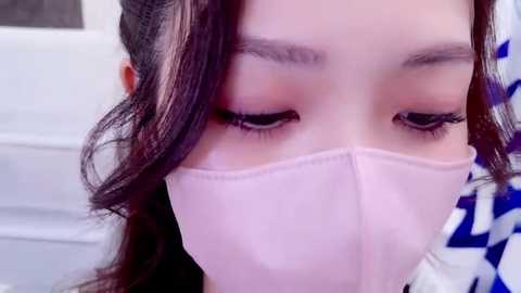 Media: Video of a woman with long brown hair, wearing a light pink face mask, and a blue and white patterned shirt. Her eyes are closed, focusing on the mask.