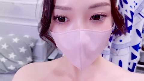 Media: A close-up video of an East Asian woman with fair skin, dark hair, and wearing a pink face mask, lying on a bed with white and blue patterned sheets.