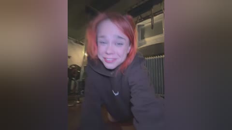 Media: A video of a young woman with red hair, wearing a black hoodie, and holding a camera in a dimly lit room with kitchen appliances and a radiator.