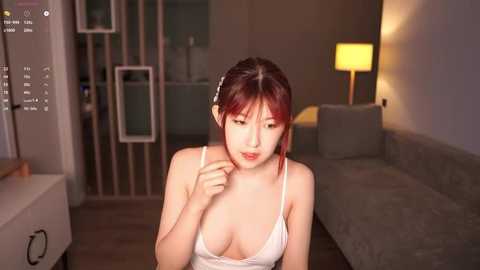 Media: A video of an East Asian woman with fair skin, red hair, and a slender physique, wearing a white lace bra, sitting in a modern, dimly-lit living room with a beige sofa and a yellow lamp.