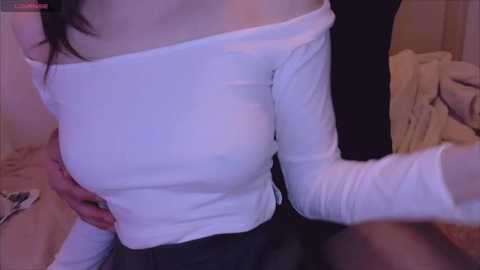 Media: Video of a woman with light skin and dark hair wearing a tight white off-shoulder top, revealing her ample breasts and a hand on her stomach. Background shows a beige blanket and a wooden floor.