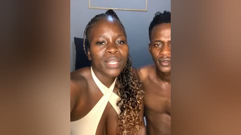 Media: Video of two shirtless black men and woman with dark skin, wearing a beige halter top, smiling, against a plain blue wall, blurred background.