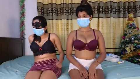 Media: Video of two South Asian women in face masks, one in black lace bra and red skirt, the other in maroon bra and white shorts, sitting on a bed, with a Christmas tree in the background.