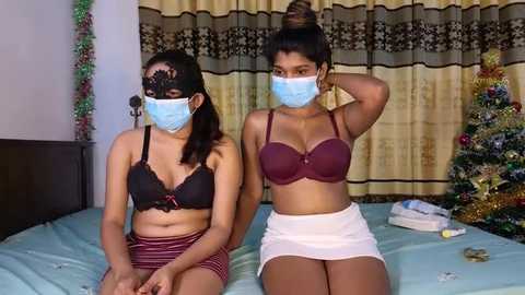 Media: Video of two young women with light brown skin, wearing face masks and festive lingerie, sitting on a bed with a Christmas tree in the background.