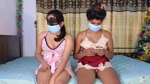 Media: Video of two women in light blue surgical masks, pink lingerie, and matching lace masks, sitting on a bed with light blue sheets, in a bedroom with brown curtains and a Christmas tree.