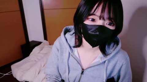 Media: A video of an East Asian woman with straight black hair, wearing a black face mask, gray hoodie, and a beige blanket. She sits on a bed in a dimly lit room with wooden paneling.