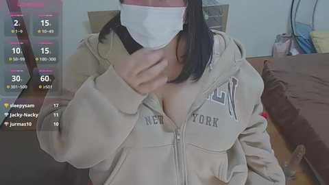 Media: A video of a person in a beige hoodie with the New York logo, wearing a white mask, and covering their mouth, standing in a bedroom with a bed and a computer screen showing game stats.