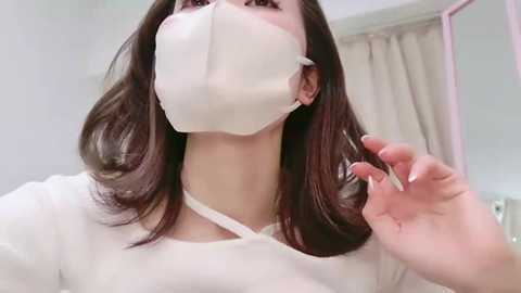 Media: A video of an Asian woman with long, straight, dark brown hair, wearing a white surgical mask, a white robe, and a white headband, brushing her hair in a bright, sterile room with a beige curtain in the background.