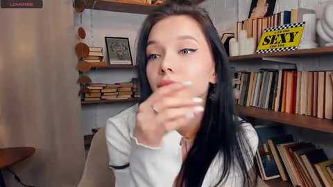 A video of a young woman with long black hair, wearing a white sweater, sitting in a book-filled room, holding a book near her mouth.