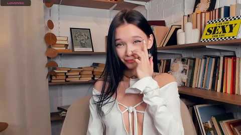 Video of a young Asian woman with long black hair, wearing a white off-shoulder blouse and a necklace, seated in a book-filled room.