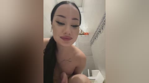 Media: A video captures a nude woman with long black hair, wearing makeup, and a tattoo on her shoulder, smiling while standing in a bathroom with white tiles and a shower curtain.