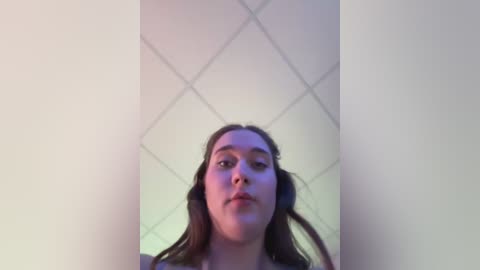 Media: Video of a young woman with long brown hair, light skin, and a neutral expression, taken from a low angle. She stands in a room with white ceiling tiles and soft lighting.