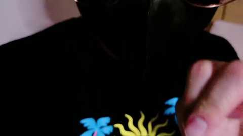 Media: A close-up video of a person wearing a black mask, black shirt with a colorful graphic, and glasses, with a hand holding a cup in the foreground.