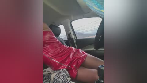 Media: Video of a woman in a red vinyl dress reclining in a car, with a blurry, colorful ceiling and a checkered blanket in the background.