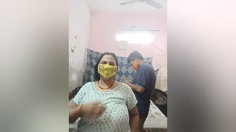 Media: A video captures a pregnant woman in a yellow mask, standing in a dimly lit, worn-out room with peeling pink walls. Behind her, a man in a dark shirt is seated.