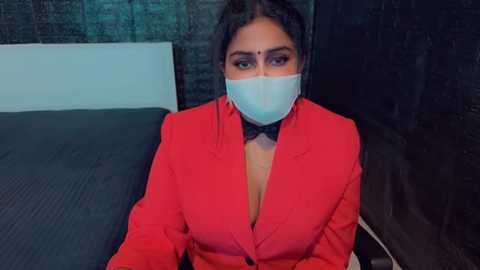 Media: Video of a South Asian woman with dark hair, wearing a red blazer, black bow tie, and a white surgical mask, seated on a dark bed in a dimly lit room.