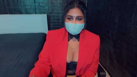 Media: Video of a woman with medium skin tone, wearing a red blazer, black bow tie, and a face mask, lying on a dark bed, with a dark brick wall as the background.