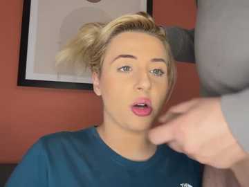 Media: Video of a blonde woman with light skin and pink lips, wearing a blue shirt, being touched by a hand in a studio with orange walls and framed artwork.