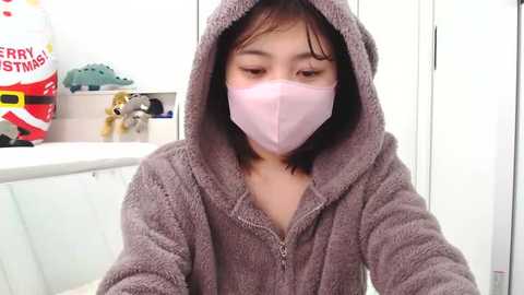 Video of an Asian woman with short black hair wearing a light pink face mask and a gray, hooded bathrobe, sitting indoors near a white sink with a \"Merry Christmas\" sign and a plush toy.