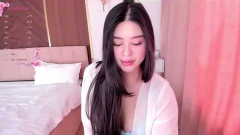 Media: Video of a young Asian woman with long black hair, wearing a white robe, sitting on a bed with white sheets and pink curtains in the background.