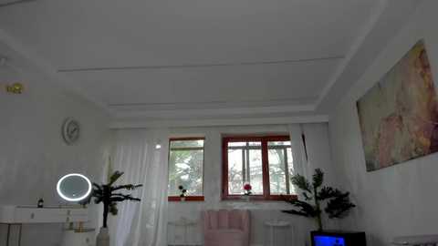 Media: Video of a modern, minimalist bedroom with white walls, a large window with sheer white curtains, a pink blanket, a round mirror with a light ring, a potted plant, and abstract art on the wall.