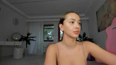 Media: Video of a light-skinned, brunette woman with a slim build, wearing a pink spaghetti-strap top, in a minimalist, brightly lit room with white walls, a round mirror, and a window with greenery outside.