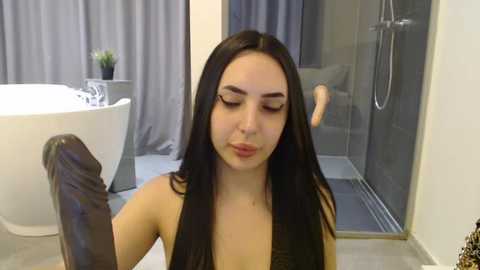 Media: Video of a young woman with long black hair, fair skin, and closed eyes, holding a large, dark dildo, standing in a modern bathroom with a white bathtub, grey curtains, and a glass shower.