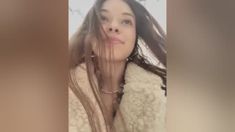 A video of a young woman with long, dark hair, wearing a beige, textured coat, looking upward with a neutral expression. The background is blurred.