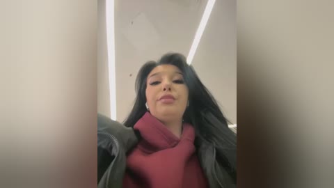 Media: Video of a young woman with long black hair, wearing a red scarf and dark jacket, standing indoors with a modern, angular ceiling and bright lights.