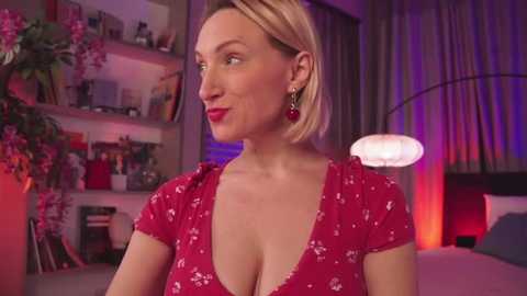 Media: Video of a blonde woman with a V-neck red dress, standing in a dimly lit room with shelves, pink flowers, and a round lamp in the background.