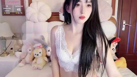 Media: Video of a fair-skinned, young Asian woman with long black hair, wearing a white lace bra, sitting on a bed. Background includes stuffed toys, a digital clock, and a wooden door.