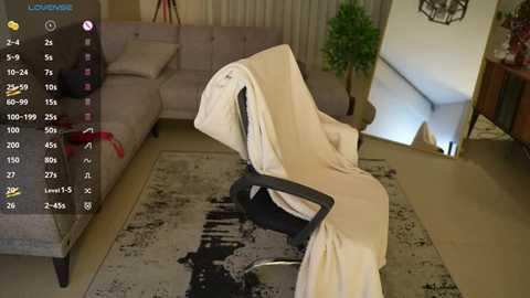 Media: Video of a modern living room with a grey couch, beige curtains, and a black office chair covered in white sheets, with a digital overlay displaying a game interface.