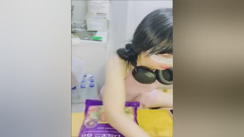 Media: Video of an East Asian woman with dark hair, wearing a pink tank top, headphones, and glasses, looking down at a purple grocery bag. Background includes a white fridge and various items.