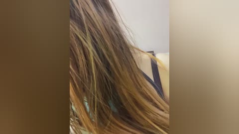 Media: Video of a woman with long, straight, light brown hair, wearing a black tank top, seen from the side. The background is a blurred, beige wall.