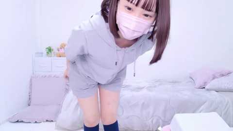 Media: A video of an Asian woman in a light gray tracksuit, kneeling with her face partially obscured by a pink face mask, in a minimalist, pastel-colored bedroom.