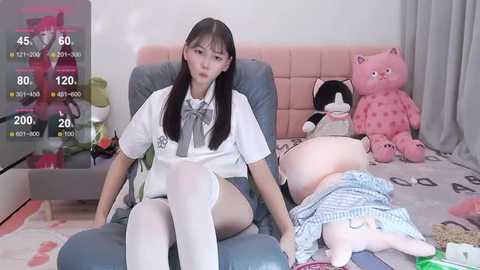 Media: A video of an East Asian woman with long black hair, wearing a white school uniform, sitting on a blue chair in a cluttered bedroom with stuffed animals and a plush teddy bear.