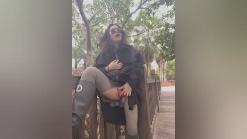 Media: Video of a woman in a black coat, sunglasses, and knee-high socks, sitting provocatively on a wooden bench in a park, legs spread, hand on her chest.