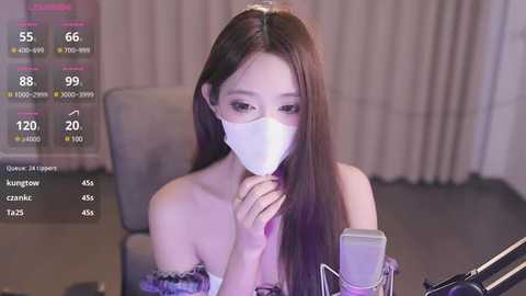 Media: Video of an East Asian woman with long brown hair, wearing a white face mask, sitting at a desk, holding a microphone. Background shows a dimly lit room with beige curtains.