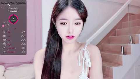 Video of an Asian woman with long, straight black hair, fair skin, and pink lipstick, wearing a white sleeveless top, standing in a modern, light-toned room with a pink wall and white staircase.