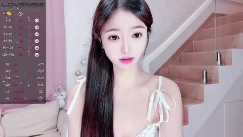 Media: Video of a young Asian woman with long black hair, fair skin, and pink lipstick, wearing a white lace bra, in a modern, pink-toned room with a beige couch and a staircase.