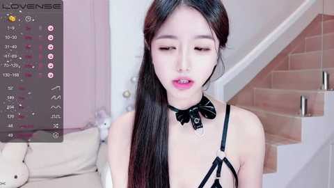 Media: Video of an Asian woman with long black hair, wearing a black leather harness and choker, standing in a modern, pastel-toned living room with plush toys and a staircase.