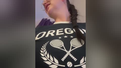 Media: Video of a woman with light skin, dark hair in a braid, wearing a black T-shirt with \"OREGON\" and tennis racket emblem. Background is blurred, indoor setting.