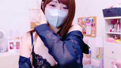 Media: Video of a young Asian woman in a dark room, wearing a silver face mask, black lace bra, and a sheer navy blue robe, with a messy background featuring a white desk, shelf, and framed pictures.