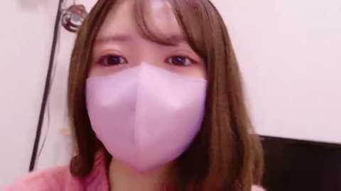 Media: Video of a young Asian woman with light skin, brown hair, and brown eyes, wearing a pink face mask, pink top, and a white wall in the background.