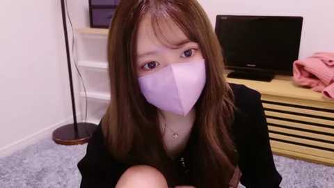 Media: Video of an Asian woman with long brown hair and light skin, wearing a pink face mask, black top, and a necklace, in a modern, light-colored living room with a flat-screen TV, wooden furniture, and a pink cloth.