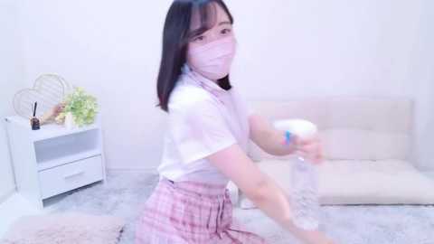 Media: Video of an East Asian woman with straight black hair, wearing a pink checkered skirt, white top, and mask, holding a white controller in a minimalist, bright living room.