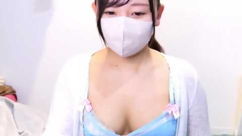 Media: A video of an East Asian woman in a white mask, light blue bra, and white cardigan, with medium breasts, sitting in a white room.