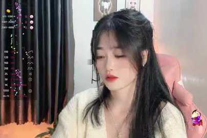 Media: A video of an East Asian woman with long black hair, fair skin, and red lipstick, wearing a white cardigan, sitting in a pink chair, indoors with a black curtain in the background.