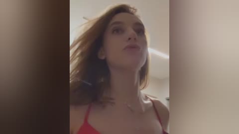 Media: Video of a young woman with shoulder-length auburn hair, wearing a red spaghetti-strap top, captured from a low angle, with soft lighting and blurred background.