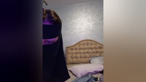 Media: A video captures a partially obscured person in a black mask, standing in a dimly lit bedroom with a tufted headboard, a bed with a light blue pillow, and a textured gray wall.